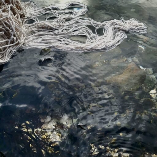 Close-up of white textile strands partially submerged in clear coastal water, with rocks, seaweed, and rippling reflections beneath the surface
